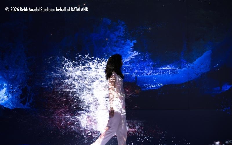 A person walks through DATALAND, surrounded by an immersive digital installation of vivid blue and white abstract visuals projected across the walls and their clothing.