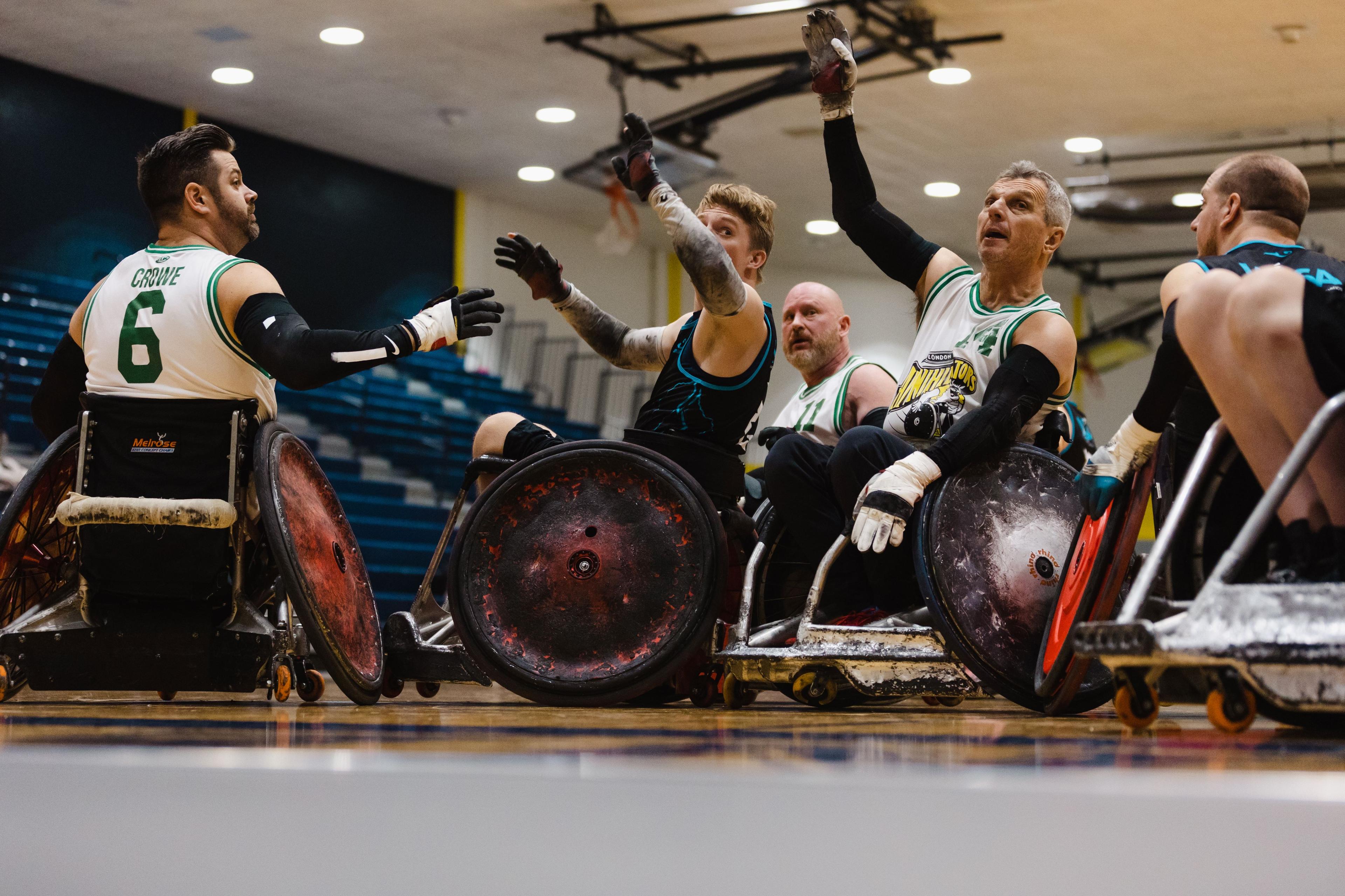 A photo of two wheelchair rugby athletes looking up at the ball with hands up trying to catch it