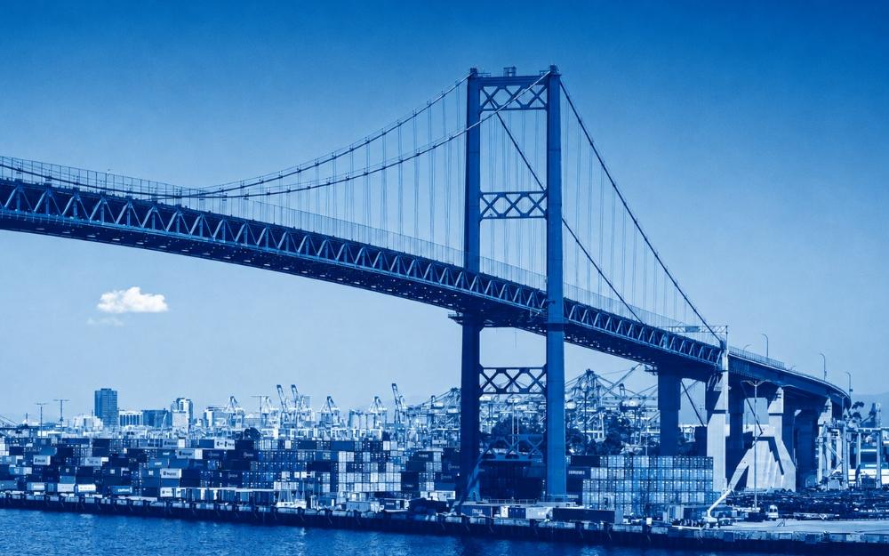 The Vincent Thomas Bridge spans the Port of Los Angeles, with stacks of shipping containers, cargo cranes, and waterfront infrastructure visible below against a clear blue sky.
