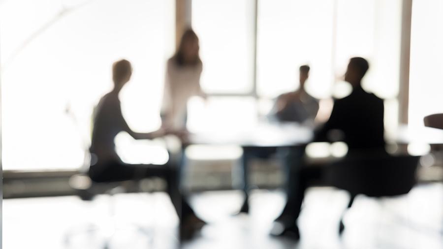 Blurred silhouettes of people seated around a table in a meeting, backlit by large windows
