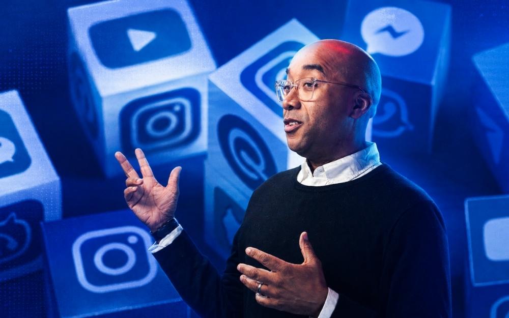 A man gestures while speaking, set against a stylized blue background featuring oversized social media icons, including Instagram, YouTube, Messenger, and other digital platforms.
