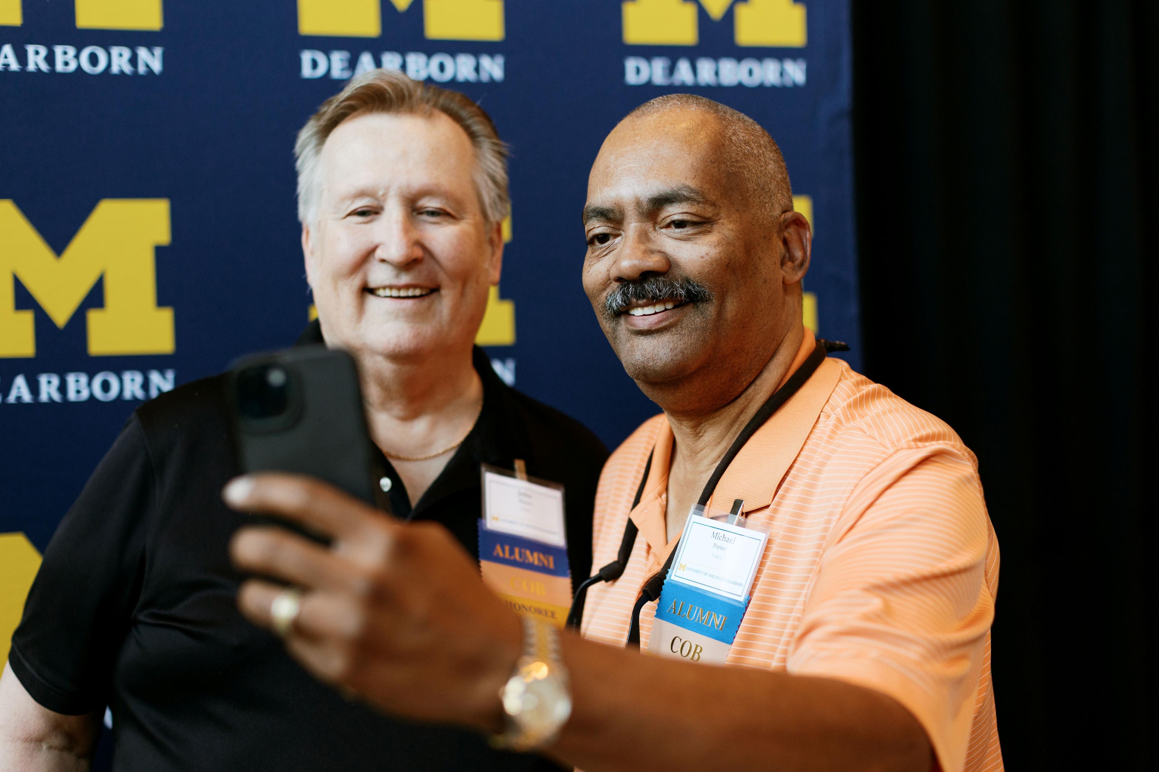 Two attendees at the 50th Jubilee Celebration pose for a selfie photo with the UM-Dearborn logo behind them.